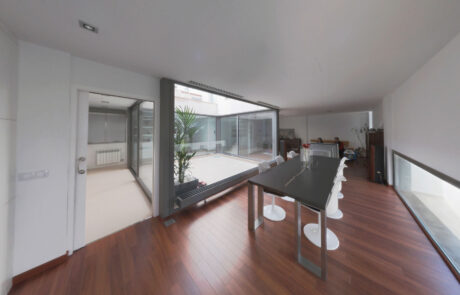 comedor de una vivienda donde se ve una mesa con sillas y al fondo dos personas sentadas en un sofá viendo la tele