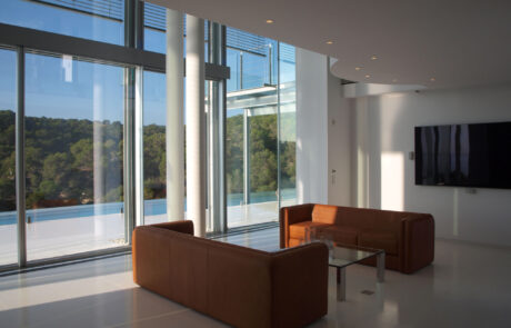 interior de una vivienda, se ven dos sofás, una mesa y una tele visión al fondo en la pared. Pared acristalada con vistas al mar